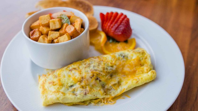 The image shows an omelet on a plate, served with a bowl of diced potatoes, sliced strawberries, orange slices, and pancakes.