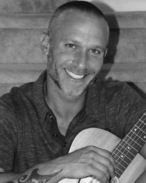 A person with a tattoo, smiling and holding an acoustic guitar, sitting on a staircase.