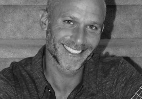 A person with a tattoo, smiling and holding an acoustic guitar, sitting on a staircase.