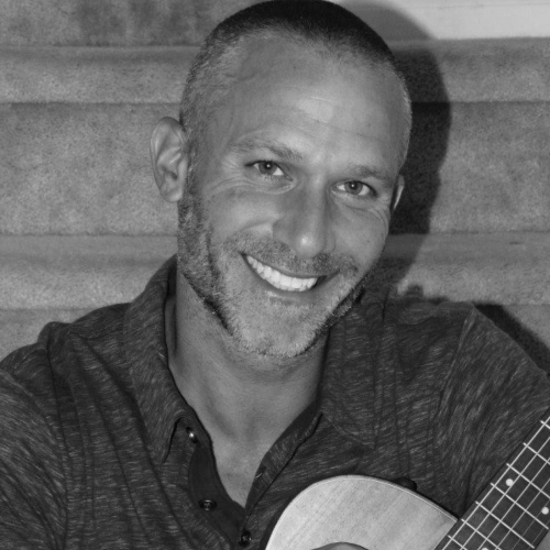 A person with a tattoo, smiling and holding an acoustic guitar, sitting on a staircase.