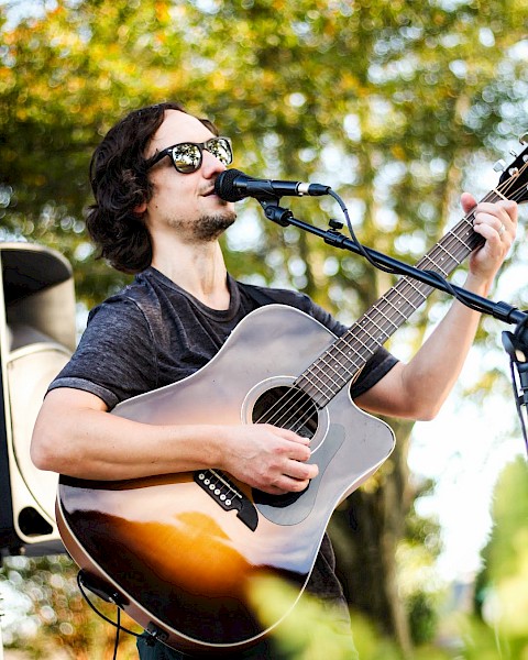 A person is playing an acoustic guitar and singing outdoors, surrounded by greenery, wearing sunglasses.