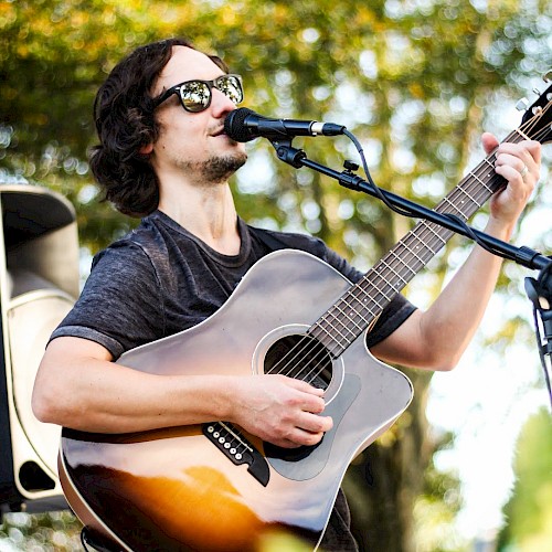 A person is playing an acoustic guitar and singing outdoors, surrounded by greenery, wearing sunglasses.