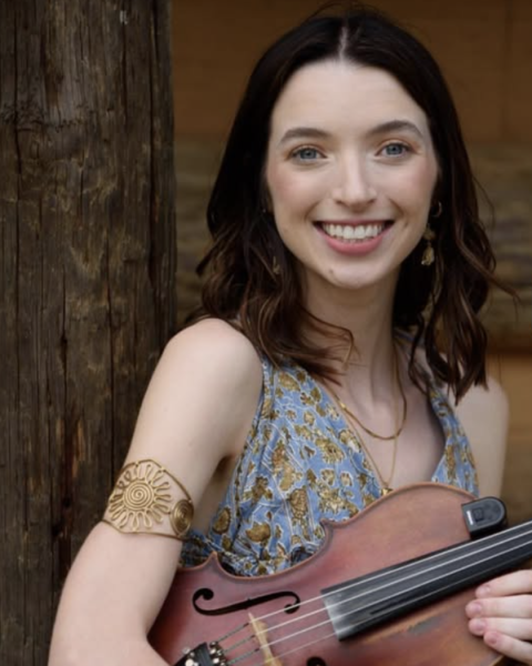 A person is smiling while holding a violin, standing by a wooden post, wearing a patterned dress and a gold armband.