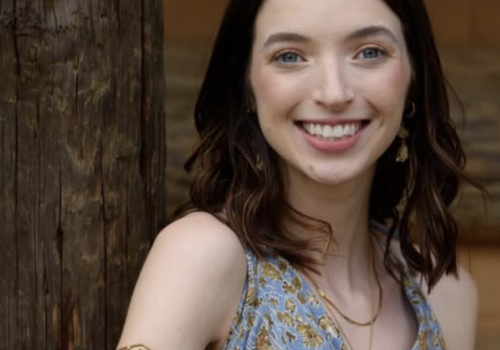 A person is smiling while holding a violin, standing by a wooden post, wearing a patterned dress and a gold armband.