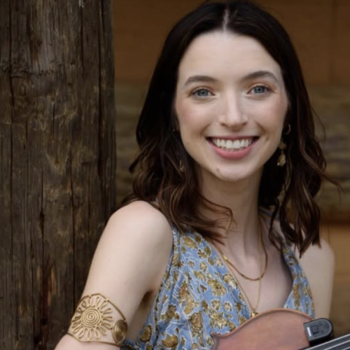 A person is smiling while holding a violin, standing by a wooden post, wearing a patterned dress and a gold armband.