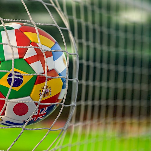 A soccer ball with national flags, including Portugal, Brazil, Japan, and the UK, caught in a goal net on a field.