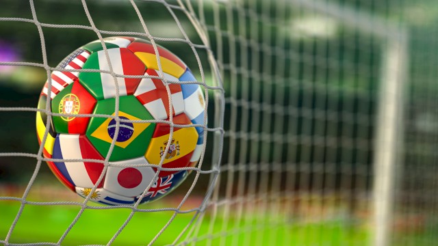 A soccer ball with national flags, including Portugal, Brazil, Japan, and the UK, caught in a goal net on a field.