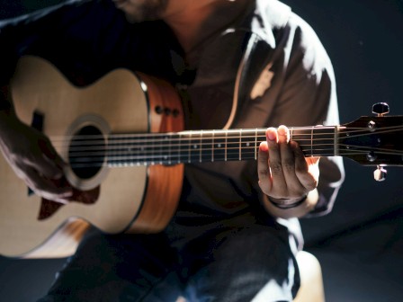 A person is playing an acoustic guitar in a dimly lit setting, focusing on the fretboard with soft light illuminating their hands and instrument.