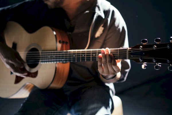 A person is playing an acoustic guitar in a dimly lit setting, focusing on the fretboard with soft light illuminating their hands and instrument.