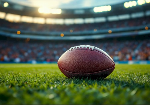 A close-up of an American football resting on the grass on a football field with stadium lights and fans in the blurred background.
