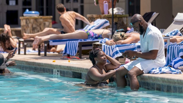 People relax by the poolside, with some chatting, sunbathing, and using their phones in a lively poolside scene.
