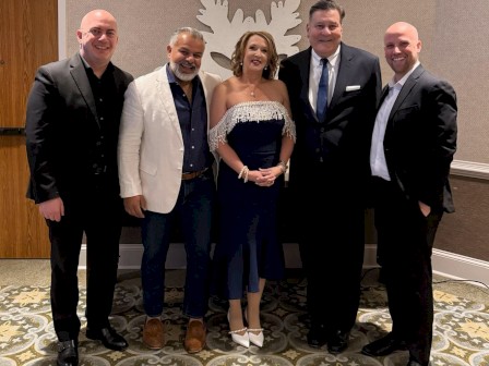 A group of five people dressed formally, standing together in a room with patterned carpet and a decorative wall behind them.