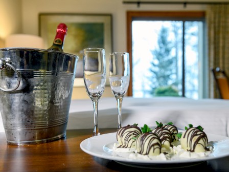 The image shows a metal ice bucket with a bottle, two empty wine glasses, and a plate of chocolate-covered strawberries by a bed.