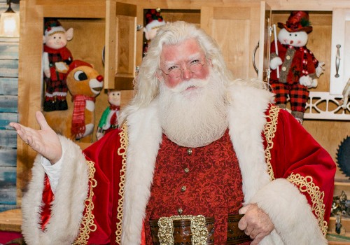 A person dressed as Santa Claus stands indoors, wearing a red and white coat with toys and festive decorations in the background.