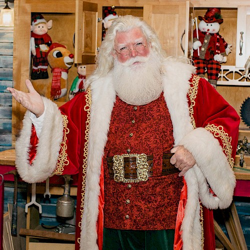 A person dressed as Santa Claus stands indoors, wearing a red and white coat with toys and festive decorations in the background.
