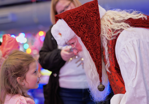 Santa in festive attire leans down to speak with a young girl during a holiday event filled with colorful lights and joyful faces.
