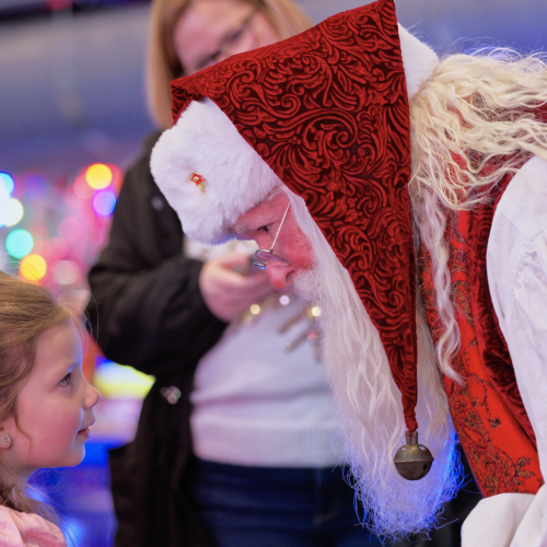 Santa in festive attire leans down to speak with a young girl during a holiday event filled with colorful lights and joyful faces.