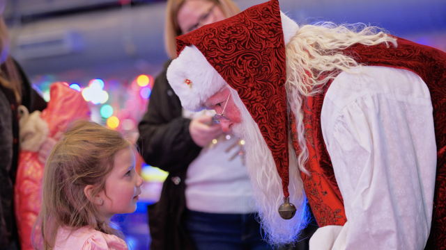 Santa in festive attire leans down to speak with a young girl during a holiday event filled with colorful lights and joyful faces.