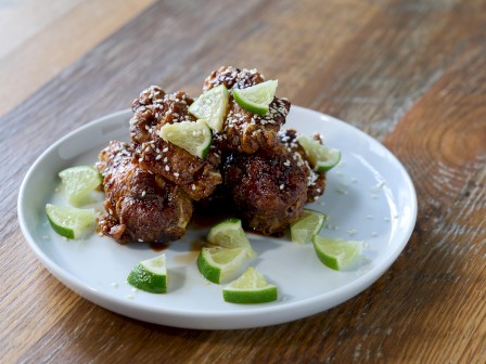 The image shows a plate of glazed chicken wings garnished with lime wedges, sprinkled with sesame seeds on a wooden table.