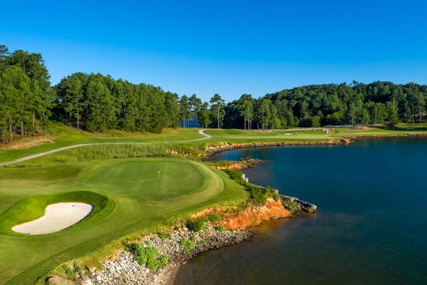 A scenic golf course with a green, bunker, and a shoreline next to a lake; surrounded by lush trees under a clear blue sky.