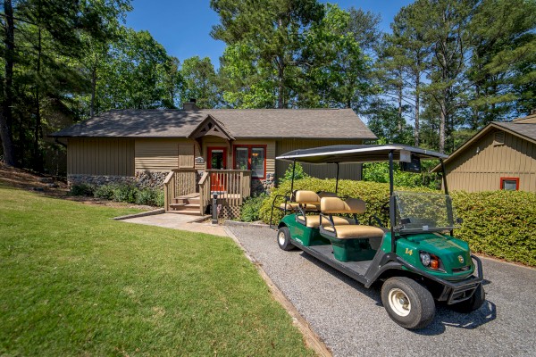 A small cabin with a porch is surrounded by trees and greenery, and a golf cart is parked on the path leading to it.