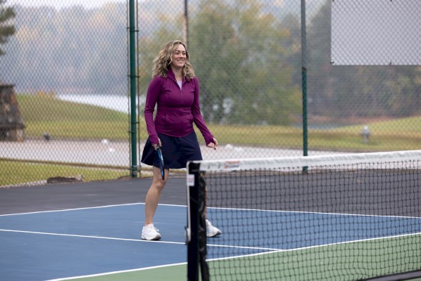 A person is standing on a tennis court with a racquet in hand, dressed in athletic wear, and surrounded by a green outdoor setting.