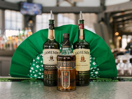 Three whiskey bottles are displayed on a bar counter with a decorative green backdrop in a bar setting.