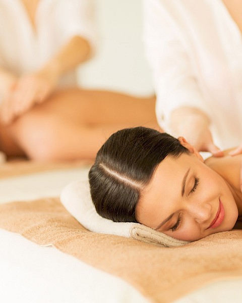 Two people are receiving a relaxing massage while lying on spa beds, with therapists attending to them in a calm environment.