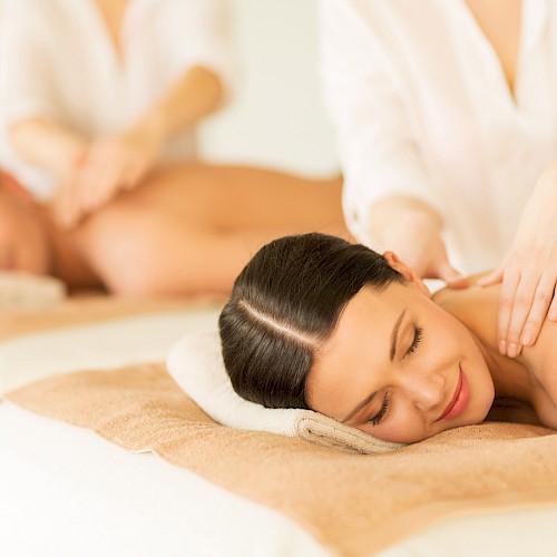 Two people are receiving a relaxing massage while lying on spa beds, with therapists attending to them in a calm environment.