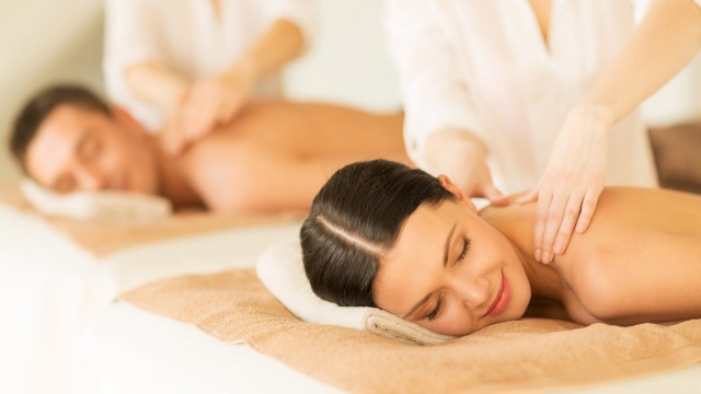 Two people are receiving a relaxing massage while lying on spa beds, with therapists attending to them in a calm environment.
