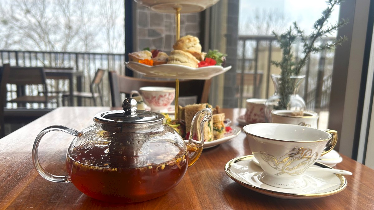 A teapot with tea, a cup, and a tiered tray of desserts and sandwiches are set on a wooden table by a window with a view.