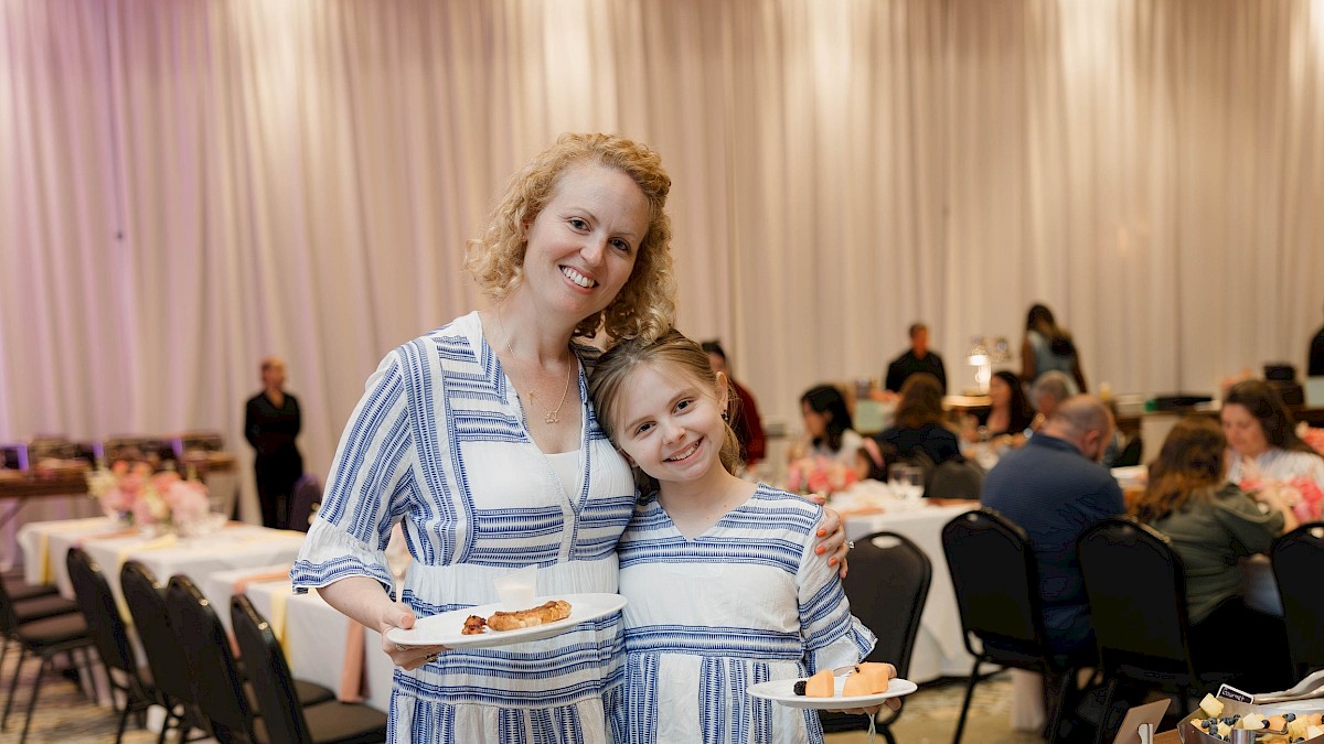 A woman and girl in matching blue and white striped dresses smile at a social event, holding plates of dessert.