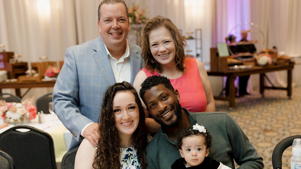 A group of people, including a baby, gathered around a table at a celebration or special event with plates, flowers, and drinks.