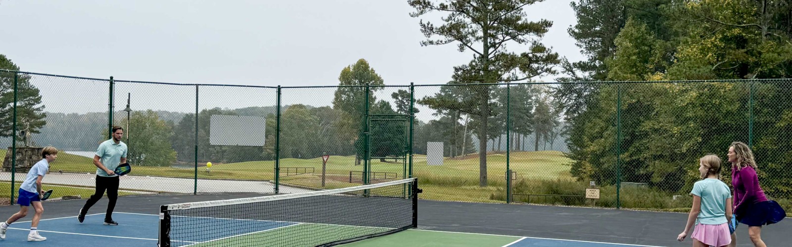 Four people playing pickleball on an outdoor court surrounded by trees and a fence.