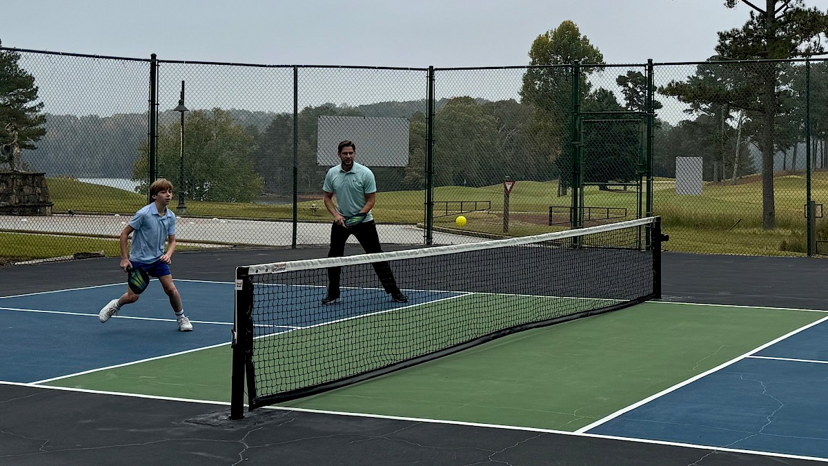 Two people are playing pickleball on an outdoor court, surrounded by a fence and trees in the background.