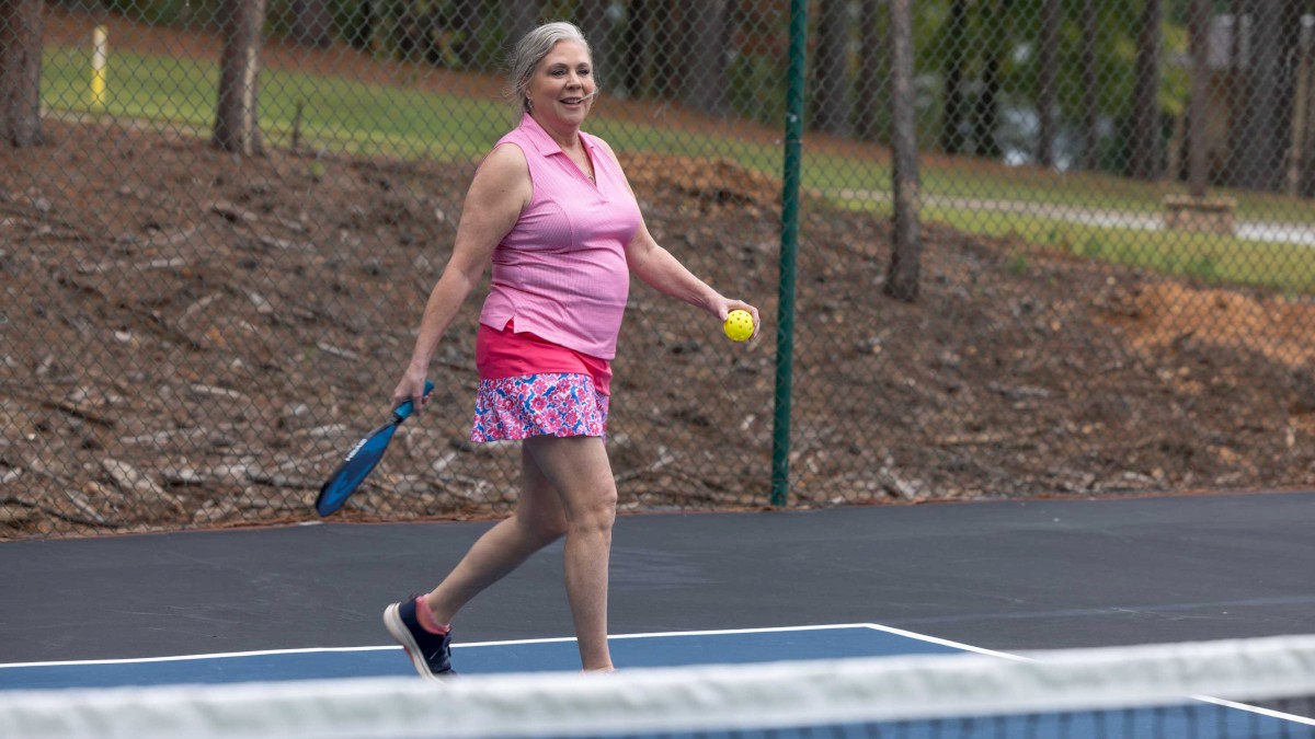 A person is playing pickleball on an outdoor court, holding a paddle and a ball, with a forest background.