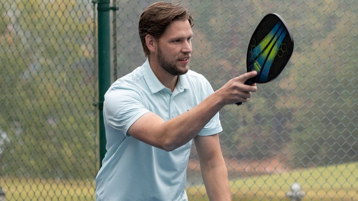 A person is playing pickleball on an outdoor court, holding a paddle and preparing to hit the ball, near a fence.