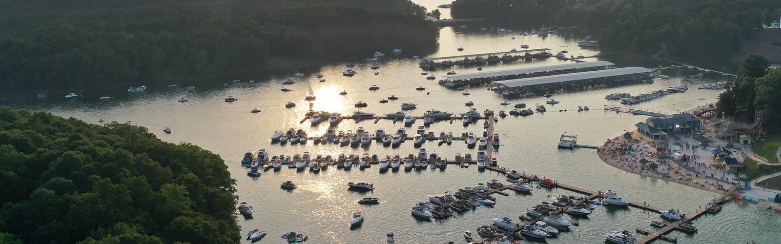 The image shows a marina filled with numerous boats docked in a calm body of water, surrounded by lush greenery and distant hills under a setting sun.