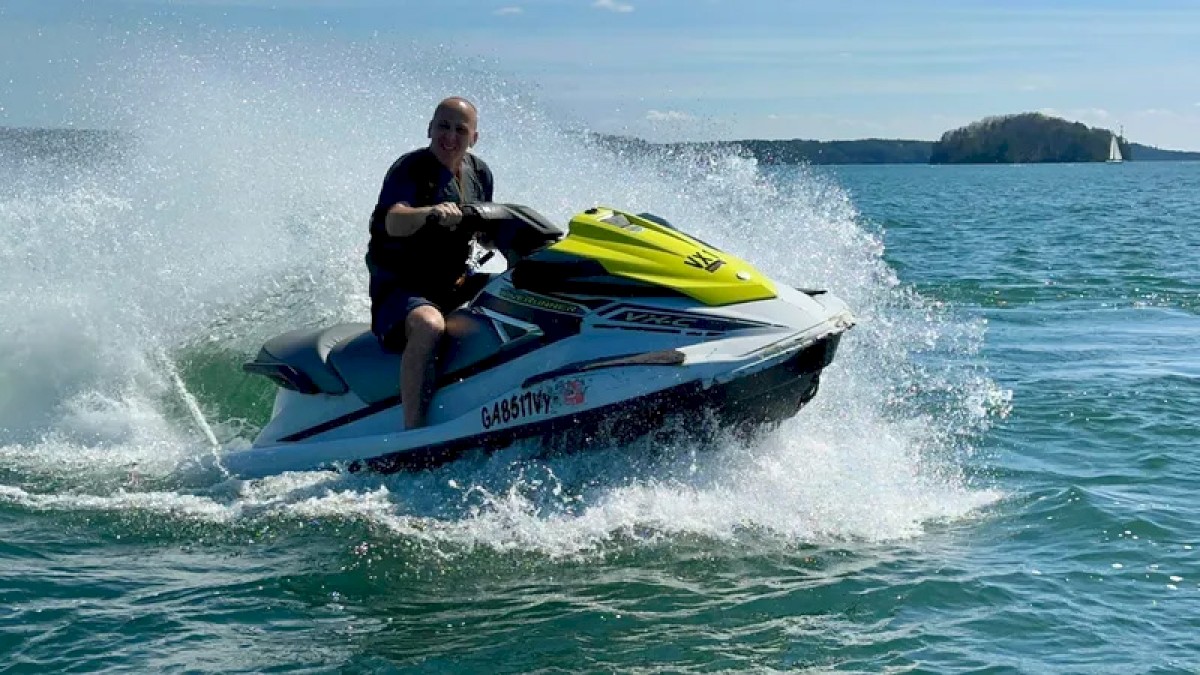 A person is riding a jet ski on a body of water, creating splashes, with a distant island and sailboat visible in the background.