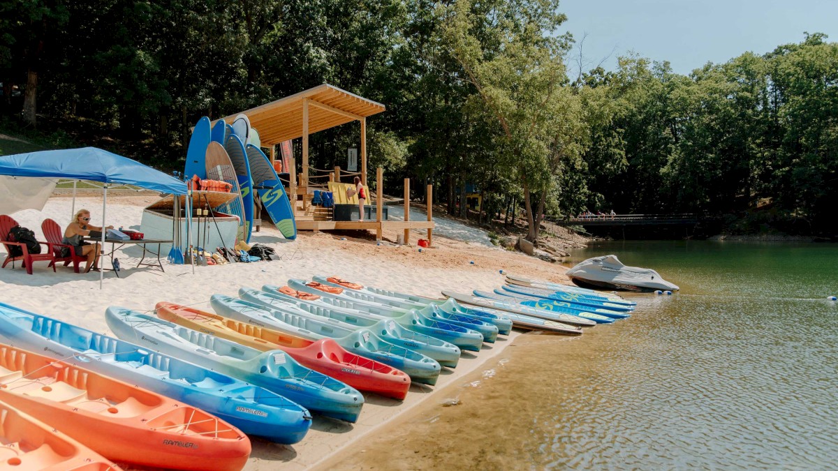 Colorful kayaks are lined up on a sandy lakeshore next to a shelter, with trees in the background and a peaceful water view.