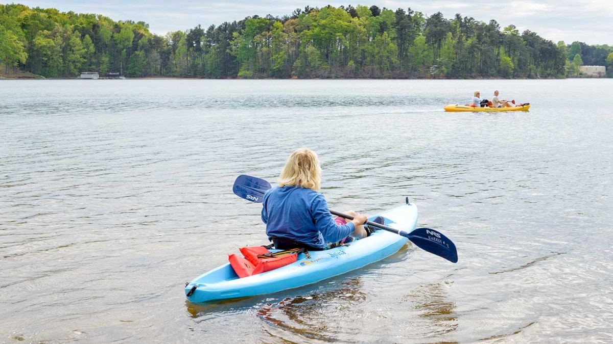 A person in a blue kayak is paddling on a lake, with another kayak and trees visible in the background.