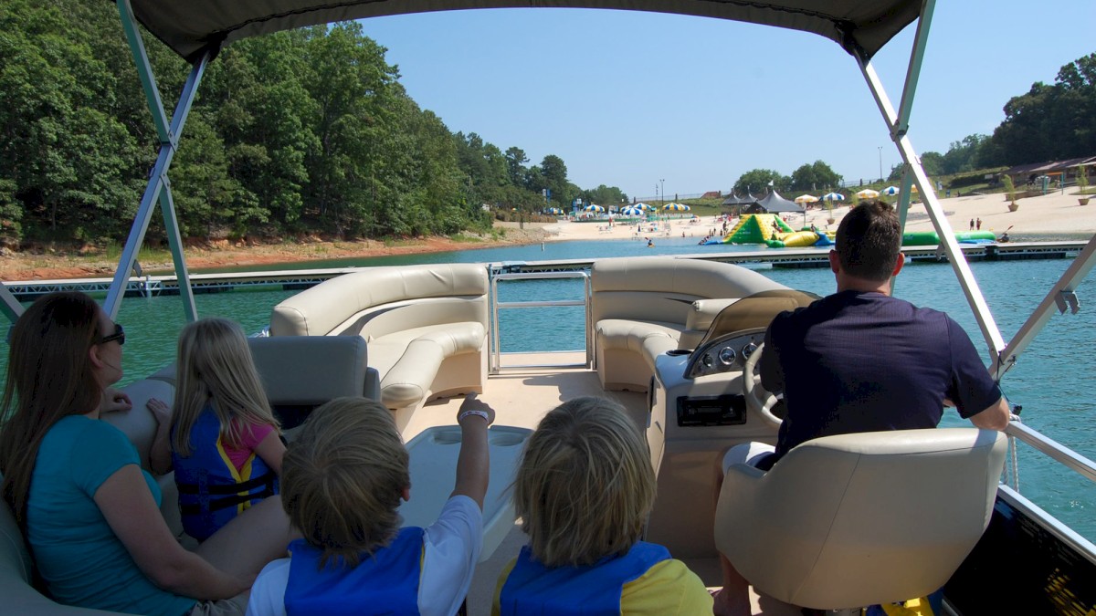 A family is on a boat ride, with the driver steering. They are heading towards a beach area with inflatables in sunny weather.