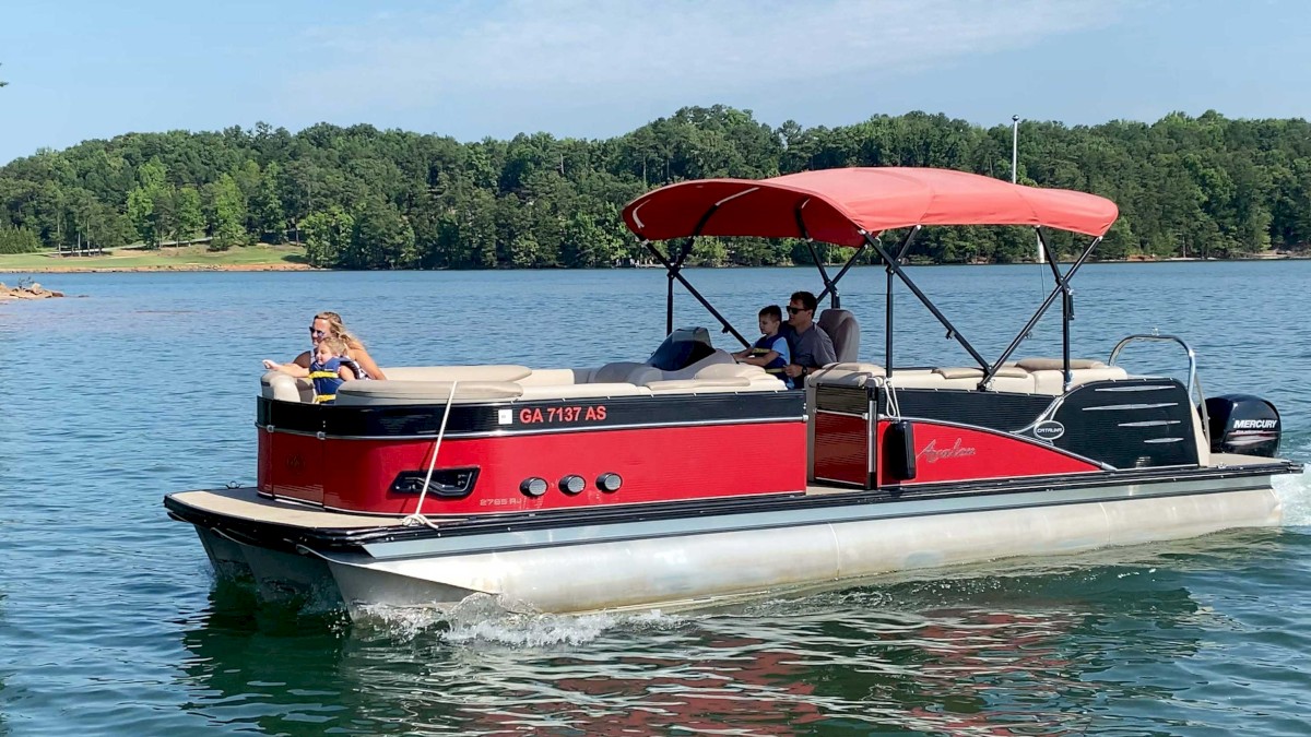 A red and black pontoon boat with a canopy is on a lake, carrying a few people, with trees in the background.
