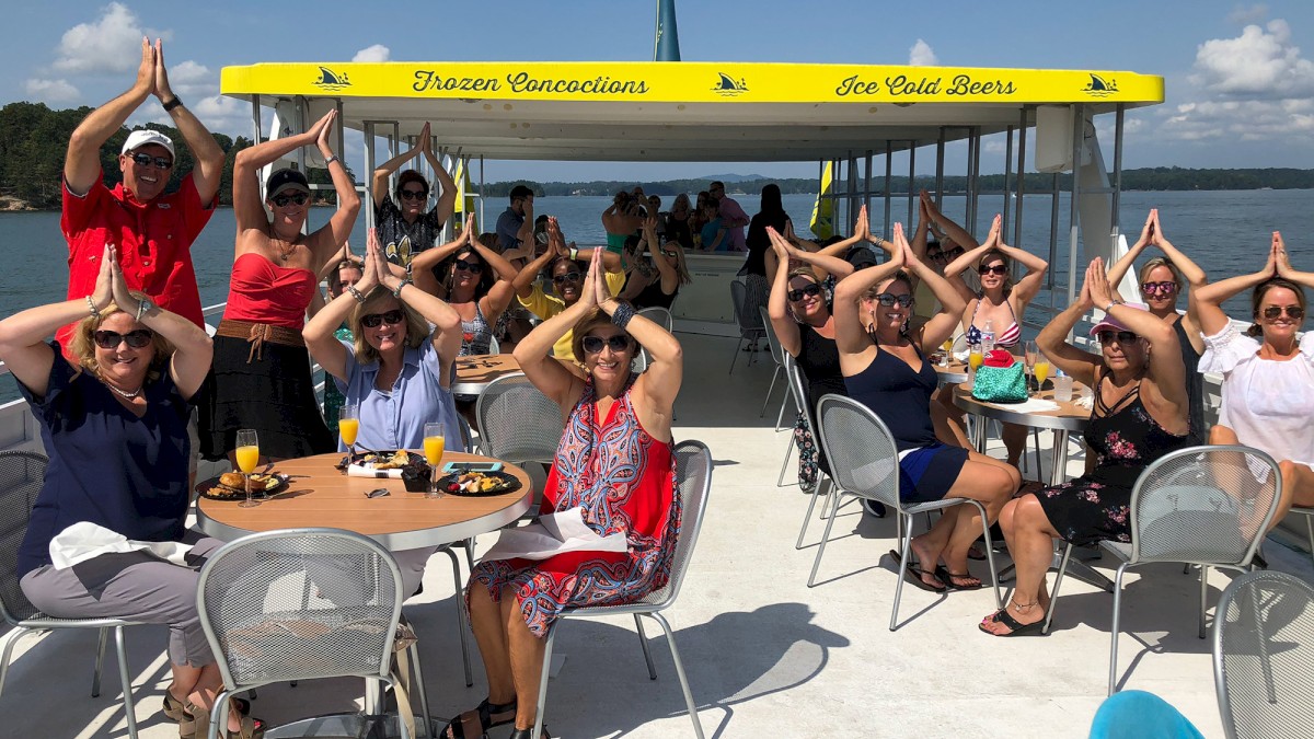 A group of people on a boat deck are posing with hands above their heads, mimicking shark fins, under a sunny sky.