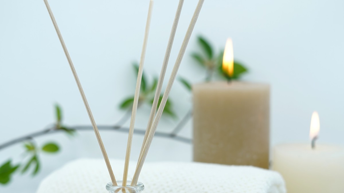 The image shows a spa setup with scented reed diffusers, rolled white towels, and lit candles, accompanied by green leaves.