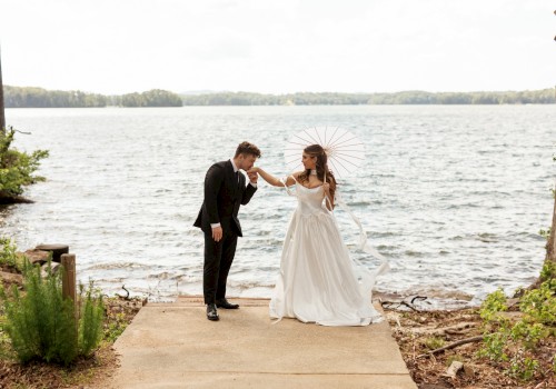 A bride and groom share a sweet moment by the water, with the bride holding a parasol and the groom kissing her hand at a lakeside.