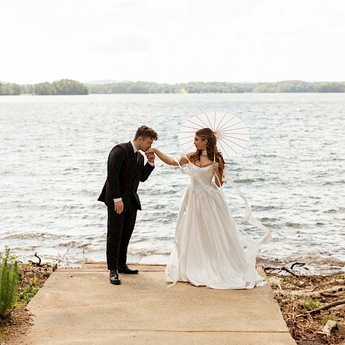 A bride and groom share a sweet moment by the water, with the bride holding a parasol and the groom kissing her hand at a lakeside.
