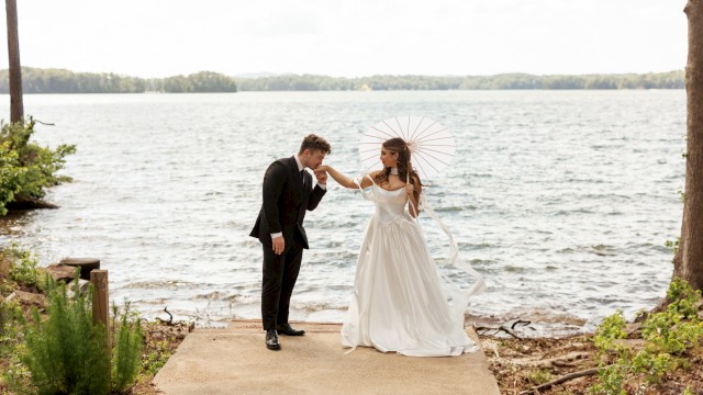 A bride and groom share a sweet moment by the water, with the bride holding a parasol and the groom kissing her hand at a lakeside.