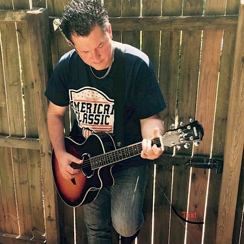 A person is playing an acoustic guitar while standing in front of a wooden fence, wearing a t-shirt and jeans.