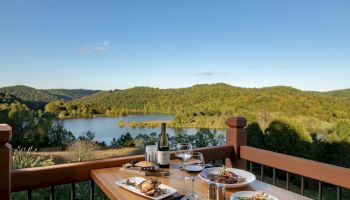 A dining table set with food and wine overlooks a serene lake surrounded by lush green hills under a clear blue sky.